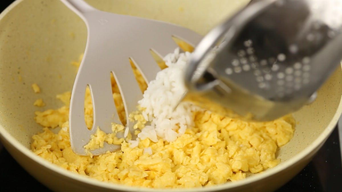 Stainless steel bowl of rice being poured into skillet with eggs