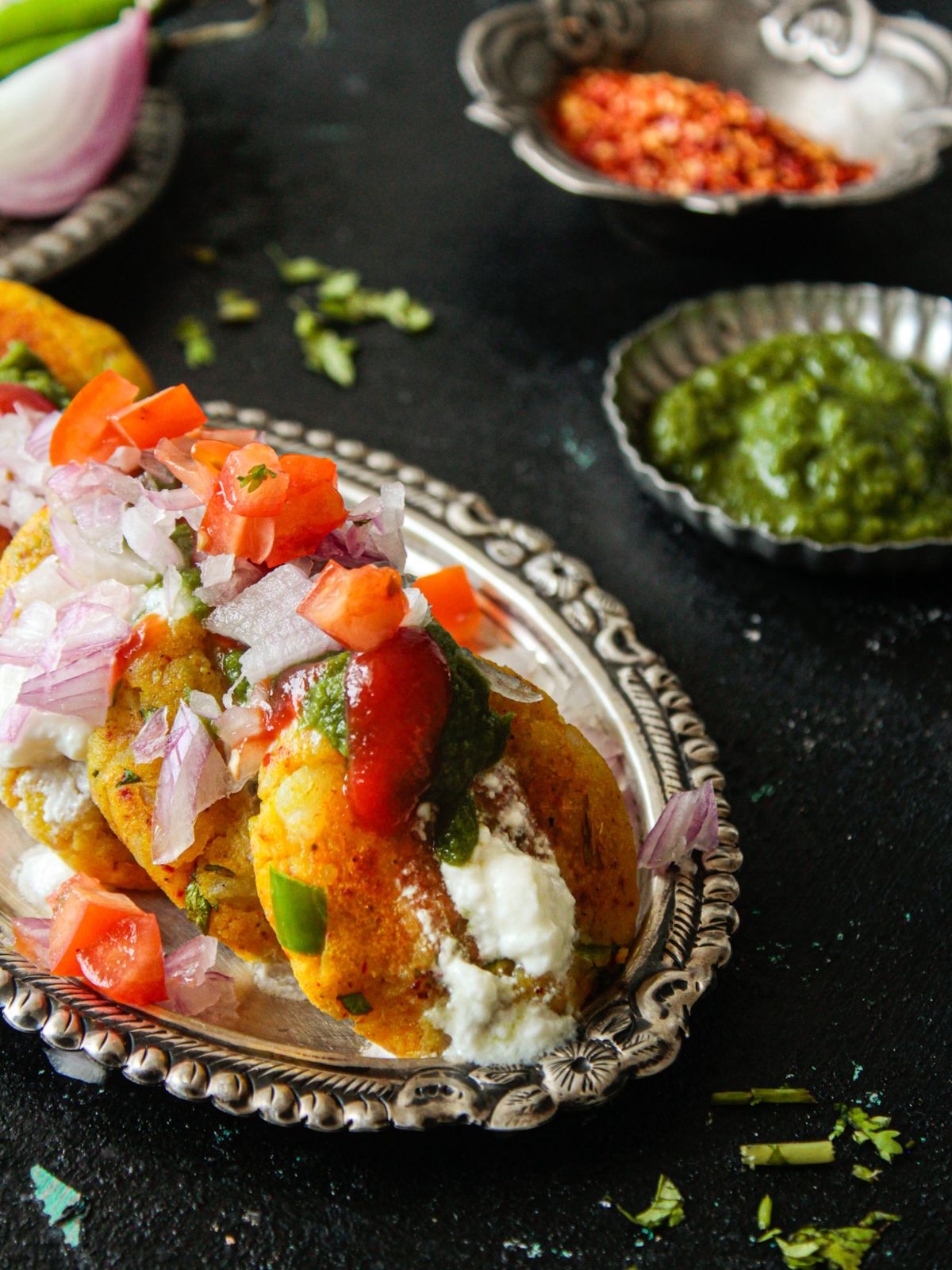 Potato patties topped with chaat on silver platter sitting on black table