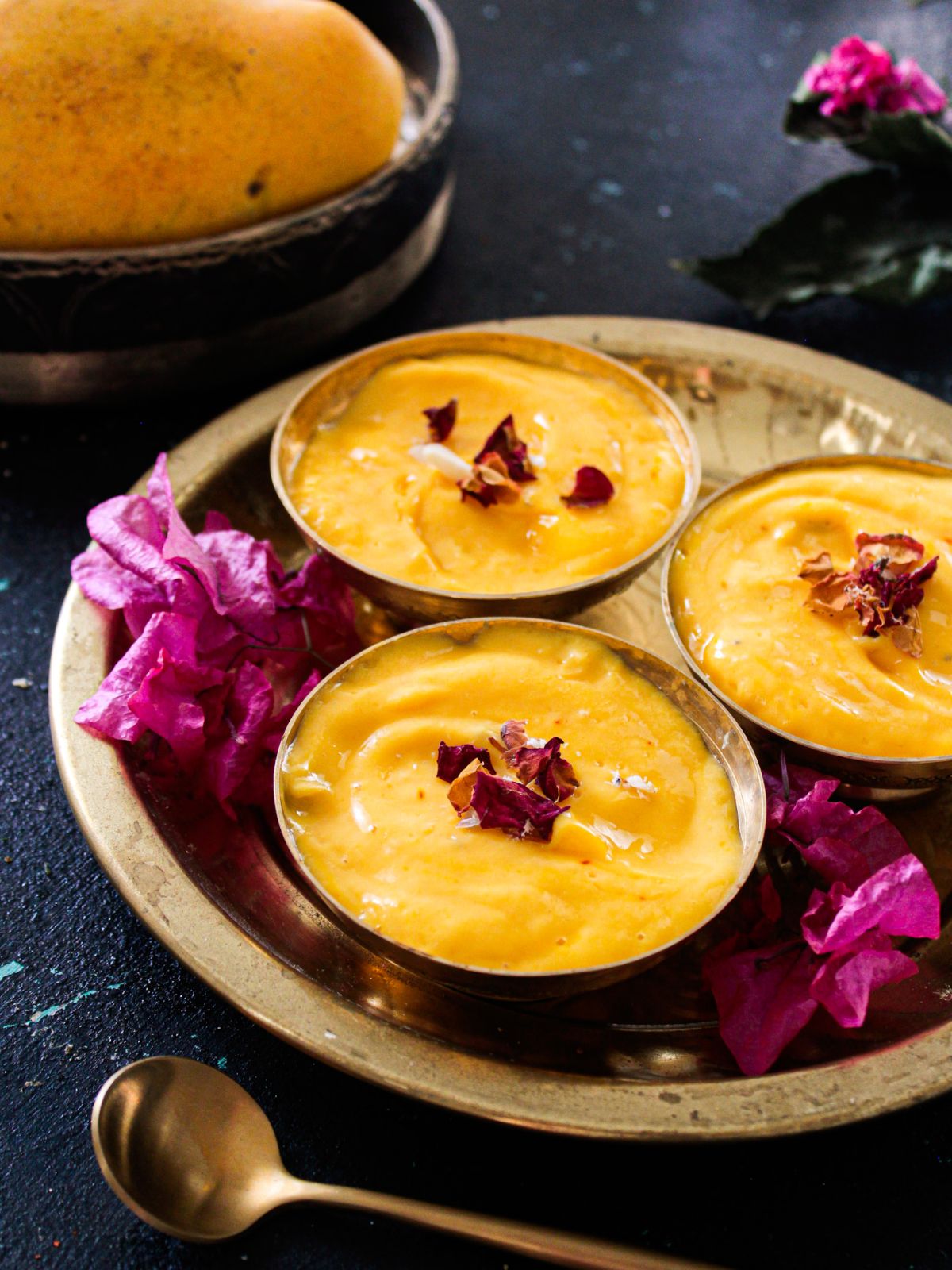 Mango shrikhand topped with rose petals in small gold bowls next to pink flowers