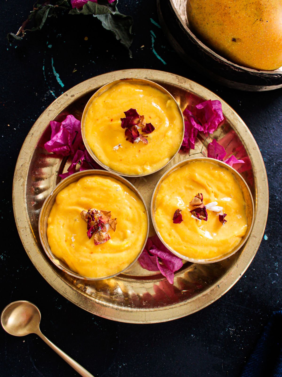 Three small gold bowls filled with mango shrikhand sitting in round gold platter on black table