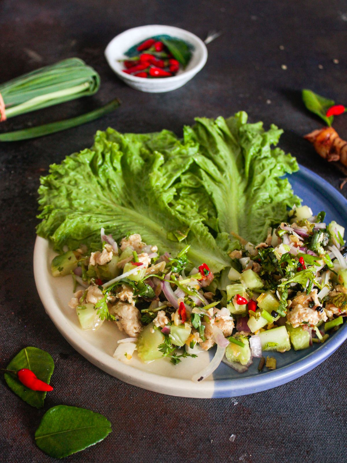 lettuce with ground chicken mixture on white and blue plate next to white bowl of red thai chiles