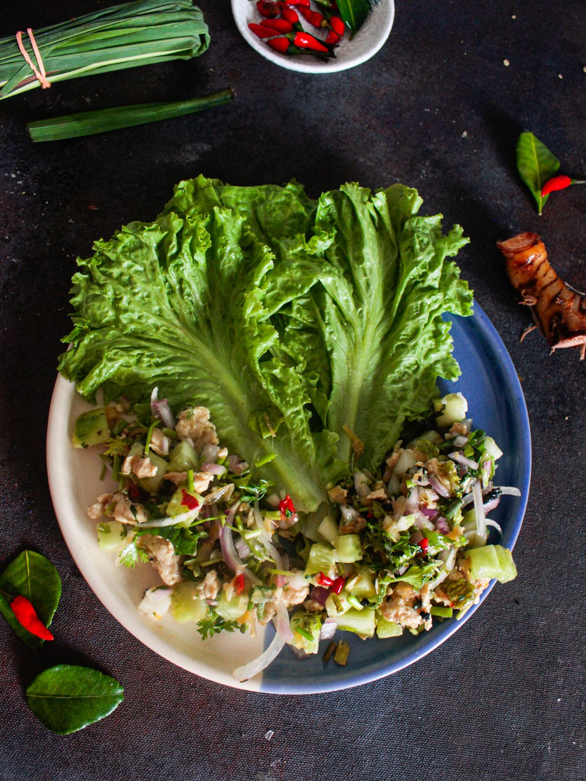 chicken larb salad on white and blue plate sitting on grey table by whole chilies